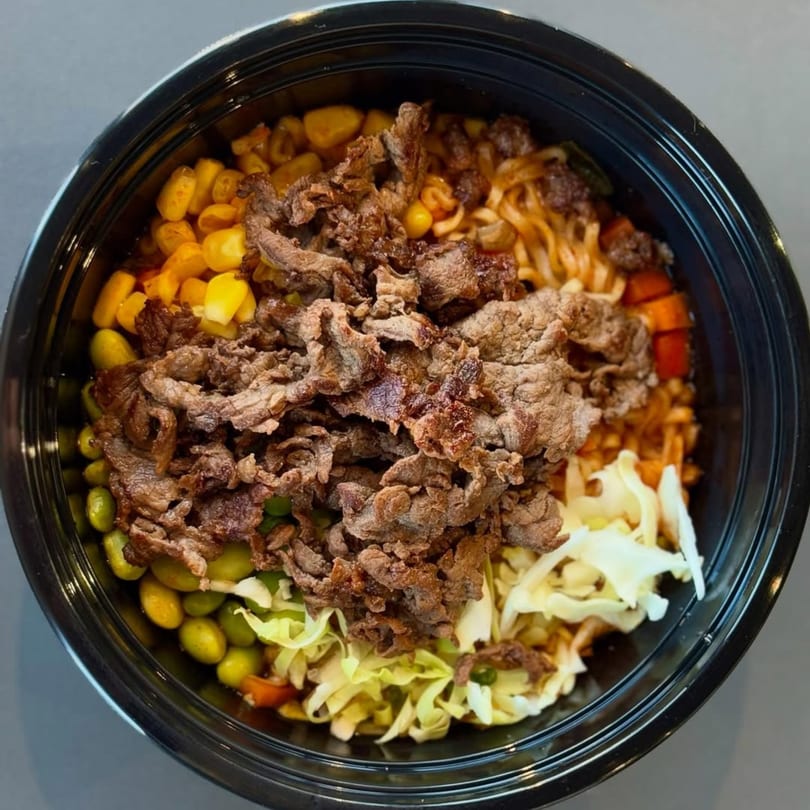 A Korean bulgogi bowl with beef, noodles, corn, edamame, and cabbage in a black bowl.