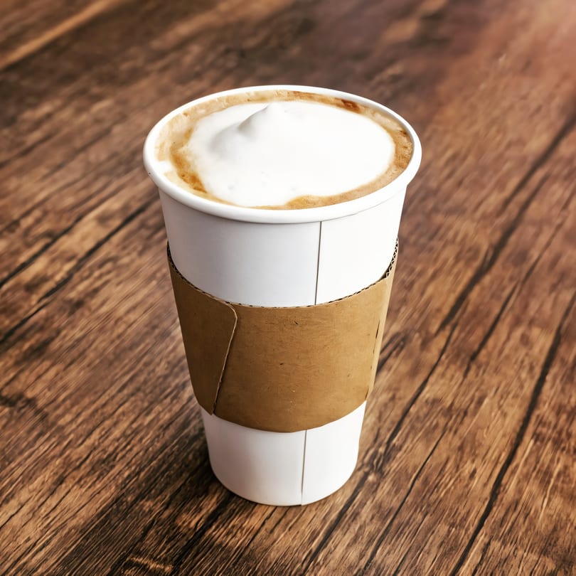 A white paper cup of coffee with foamy milk and a brown sleeve on a wooden table.