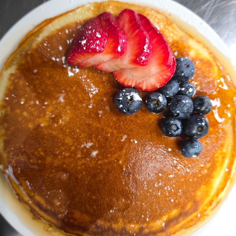 Golden pancake topped with sliced strawberries, blueberries, and powdered sugar, drizzled with syrup.